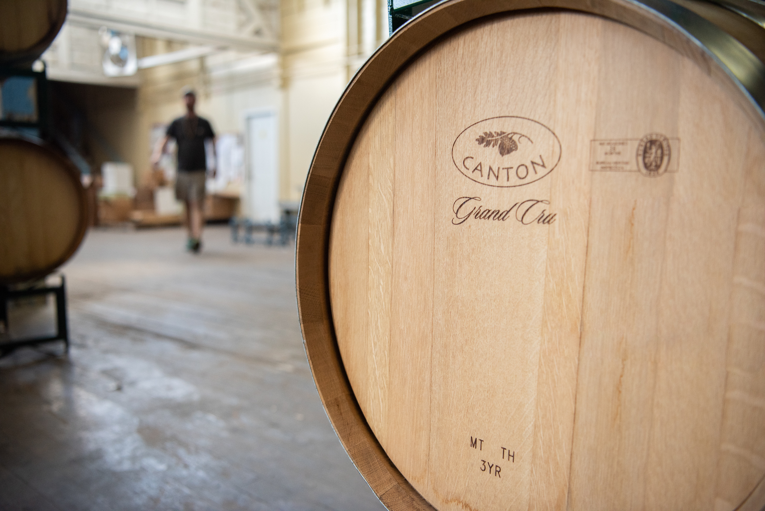 A wine barrel sits in the foreground, with other wine barrels and a person walking in the background.